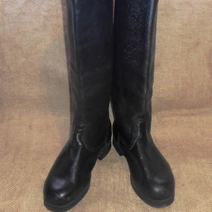 Puede incluir: Un par de botas negras de cuero hasta la rodilla. Las botas tienen una superficie texturizada y una puntera redondeada. Las botas están de pie sobre una superficie de color neutro.