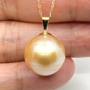 May include: A golden South Sea pearl pendant suspended from a delicate yellow gold chain. The spherical pearl features a high-luster, metallic gold surface with subtle cream undertones, secured by a polished gold bail at the top of the pendant.