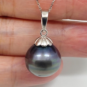 May include: A round, dark gray pearl pendant with iridescent undertones, suspended from a silver-toned metal bail and a delicate chain. The pearl features a smooth, reflective surface and is capped with a scalloped, polished silver-toned metal setting.