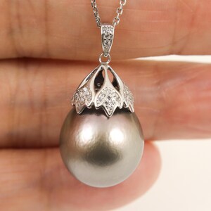 May include: A pendant necklace featuring a large, round, dark gray pearl suspended from a silver-toned metal cap. The cap is decorated with small, clear, sparkling stones arranged in a floral-like pattern, attached to a delicate silver chain.
