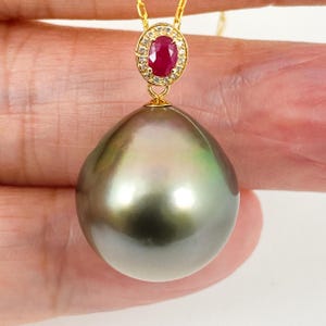 May include: A gold necklace featuring a large, pear-shaped pearl pendant. The pendant is adorned with a ruby encircled by small, clear gemstones. The pearl has a dark, lustrous surface with green and silver highlights.