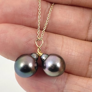 May include: A gold chain necklace featuring two dark gray-purple pearls. The pearls have a lustrous sheen and are connected by a delicate gold clasp. The necklace is held in a person's hand.