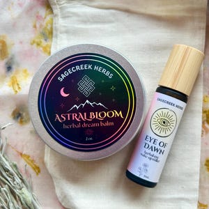 Könnte beinhalten: Ein Set aus Kräuterprodukten, darunter eine runde Dose mit der Aufschrift "Astral Bloom herbal dream balm" und eine Roll-on-Flasche mit der Aufschrift "Eye of Dawn herbal wake up roller". Die Artikel liegen auf einem cremefarbenen Beutel mit Kordelzug, mit Salbei und floralem Hintergrund.