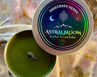 May include: A silver tin of Astral Bloom herbal dream balm, with a green balm inside. The lid features a colourful label with the text "Sagecreek Herbs" and "Astral Bloom." A sprig of lavender and a small stone are also present.