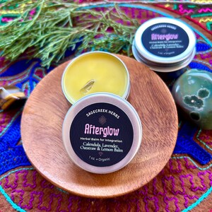 Könnte beinhalten: Auf einem Holzbrett befinden sich zwei offene Tiegel mit Kräuterbalsam und ein geschlossener Tiegel. Die Etiketten tragen die Aufschrift "Afterglow" und listen Inhaltsstoffe wie Calendula und Lavendel auf. Ein Zweig Grünzeug und ein grüner Stein sind ebenfalls zu sehen.
