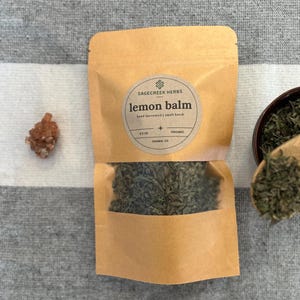 May include: A brown paper pouch with a clear window showing dried green lemon balm herbs. The pouch has a label that reads "Sagecreek Herbs lemon balm hand harvested | small batch 0.5 oz.  Prona, CO Organic".