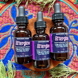 Peut inclure: Trois flacons en verre brun avec des compte-gouttes noirs, étiquetés "Afterglow" par Sagecreek Herbs. Chaque flacon contient une teinture d'intégration à base de plantes, avec une étiquette violette. Les flacons sont sur un plateau en bois.