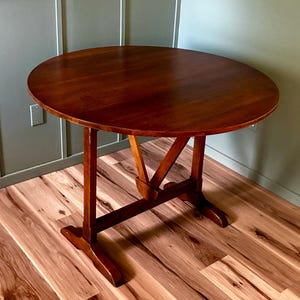 May include: A round wooden table with a smooth, dark brown finish. The table features a central trestle base with a V-shaped support structure connecting the tabletop to the horizontal base feet, providing a stable foundation for the circular top.