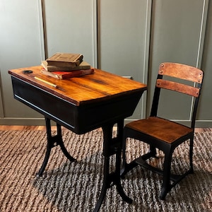 May include: A school desk and chair set featuring a wooden desktop and seat with a dark metal frame. The desk has a black storage compartment, a circular inkwell hole, and a yellow pencil. A stack of three vintage books rests on the wooden surface.