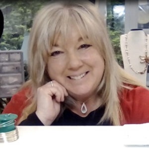 May include: A woman with blonde hair is smiling and looking at the camera. She is wearing a red shirt and a silver necklace with a teardrop pendant. She is sitting at a table with a green and silver wire spool in front of her.