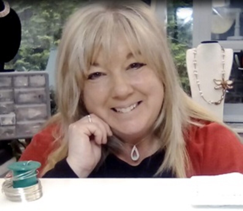 May include: A woman with blonde hair is smiling and looking at the camera. She is wearing a red shirt and a silver necklace with a teardrop pendant. She is sitting at a table with a green and silver wire spool in front of her.