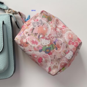 May include: A small, square, pink and white patterned pouch featuring Hello Kitty characters, bows, and hearts. The pouch is next to a light blue purse. The text "BP#5" is in the upper right corner.