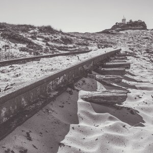 May include: A black and white photo of an abandoned set of train tracks on a sandy beach. The tracks are overgrown with weeds and the sand is blowing in the wind. The photo was taken by Wood-Bradley Photography.