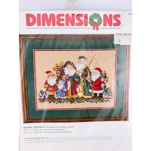 May include: A Dimensions counted cross stitch kit titled Santa Quintet designed by Barbara Mock. The packaging features a finished image of five Santas in various outfits on beige Aida fabric, framed in a dark wood frame with a green mat.
