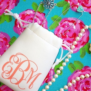 May include: A white drawstring pouch with a pink monogram "EBM" on the front. The pouch is sitting on a blue and pink floral fabric with a pearl necklace and a silver necklace with a flower pendant.