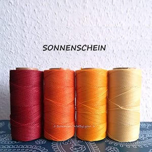 Könnte beinhalten: Vier zylindrische Garnrollen in einer Reihe, die einen Farbverlauf von tiefem Rot über leuchtendes Orange bis hin zu Goldgelb und Hellgelb aufweisen. Der Text SONNENSCHEIN steht über den Rollen und BohemianChildByLynn befindet sich darunter.