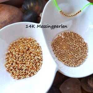 Könnte beinhalten: Eine Sammlung kleiner, runder, metallisch goldfarbener Perlen in einer weißen geteilten Schale. Ein einzelner neongrüner Faden hält eine kleine Reihe dieser Perlen. Der Text 14K Messingperlen ist in Schwarz auf der Schale aufgedruckt.