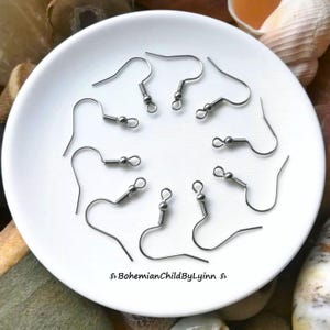 Könnte beinhalten: Eine Ansammlung von acht silberfarbenen Ohrringhaken aus Metall, die kreisförmig auf einer weißen Keramikschale angeordnet sind. Jeder Haken verfügt über ein kleines Spiralfederdetail und eine Öse am unteren Ende zur Befestigung von Schmuckteilen. Text: BohemianChildByLynn.