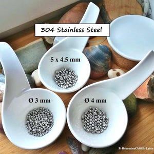 Könnte beinhalten: Drei weiße Keramikschalen gefüllt mit silbernen Metallperlen. Die Schalen sind mit den Perlengrößen beschriftet: 5 x 4,5 mm, 3 mm und 4 mm. Der Text "304 Edelstahl" steht über den Schalen.