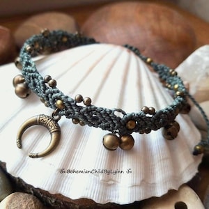 Könnte beinhalten: Ein graues Makramee-Armband mit kleinen Messingperlen und mehreren runden Messingglöckchen, die entlang des Bandes befestigt sind. Ein markanter Messing-Anhänger in Form eines Halbmonds mit geätzten geometrischen Mustern hängt in der Mitte. Text: BohemianChildByLyinn.