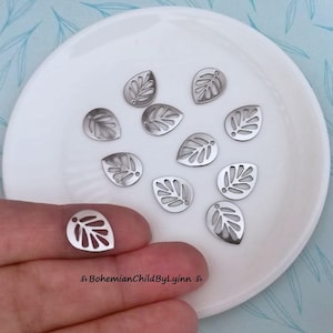 Könnte beinhalten: Ein weißer Teller mit 10 silbernen Blatt-Anhängern. Die Anhänger sind in Form eines Blattes mit einem detaillierten Design ausgeschnitten.