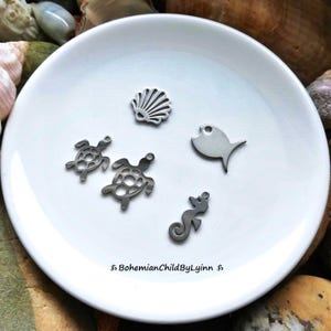 Könnte beinhalten: Ein Set aus fünf silbernen Metall-Anhängern mit einer Muschel, zwei Meeresschildkröten, einem Fisch und einem Seepferdchen. Die Anhänger befinden sich auf einem weißen Hintergrund.