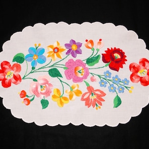 May include: Oval white embroidered table runner with a scalloped edge. The runner features a vibrant floral design with red, pink, blue, yellow, and purple flowers, and green stems. A decorative piece for a dining table.