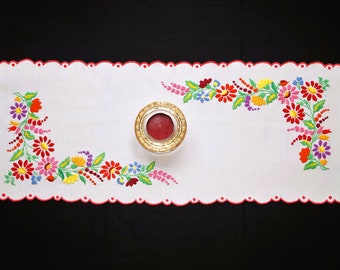 May include: White embroidered table runner with a red scalloped edge and floral designs in shades of red, pink, yellow, blue, and green. A small round candle holder with a red candle is in the center of the runner.