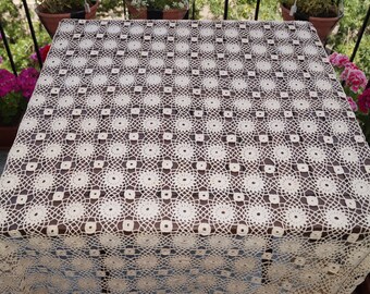 Könnte beinhalten: Eine weiße Häkeltischdecke mit einem quadratischen Muster aus zarten Blumen. Die Tischdecke liegt auf einem Holztisch.