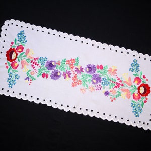 May include: White embroidered table runner with a floral design featuring pink, red, purple, and yellow flowers. The runner has a scalloped edge with small holes.