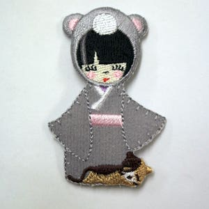 May include: Embroidered iron-on patch featuring a gray-haired girl wearing a gray hooded coat with a white circle on the hood, a pink sash, and a brown cat at her feet.