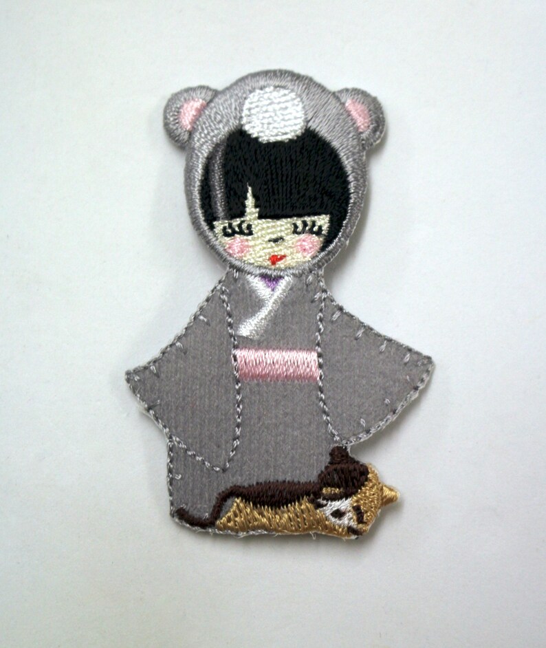 May include: Embroidered iron-on patch featuring a gray-haired girl wearing a gray hooded coat with a white circle on the hood, a pink sash, and a brown cat at her feet.