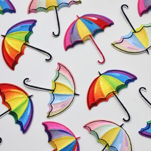May include: A collection of rainbow-coloured embroidered iron-on patches shaped like umbrellas. The patches are arranged in a scattered pattern on a white background.