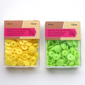 K&ouml;nnte beinhalten: Zwei Packungen mit 12 mm Kunststoff-Druckkn&ouml;pfen. Eine Packung enth&auml;lt 30 gelbe Kn&ouml;pfe und die andere 30 gr&uuml;ne Kn&ouml;pfe. Die Packungen sind mit "Boutons Pression Plastique" und "MEDIAC" beschriftet.