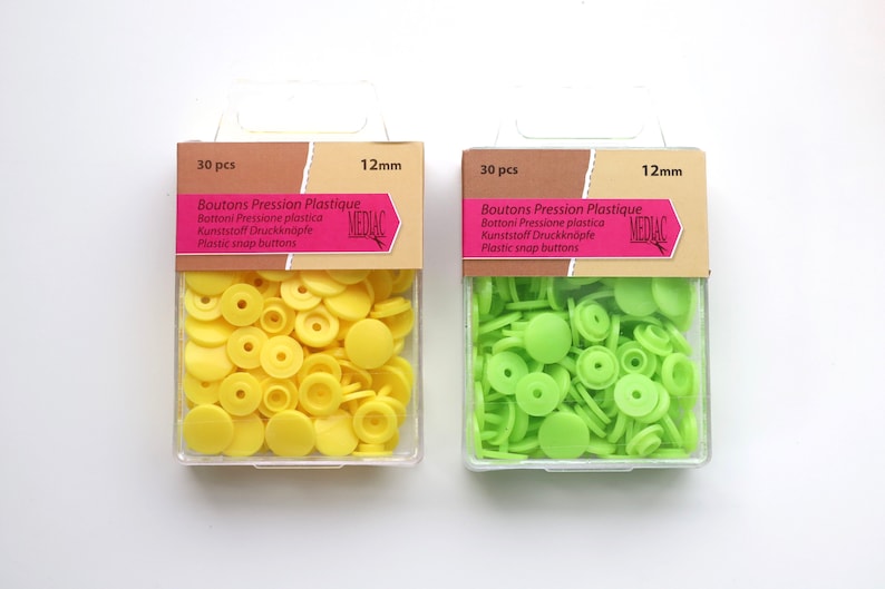 K&ouml;nnte beinhalten: Zwei Packungen mit 12 mm Kunststoff-Druckkn&ouml;pfen. Eine Packung enth&auml;lt 30 gelbe Kn&ouml;pfe und die andere 30 gr&uuml;ne Kn&ouml;pfe. Die Packungen sind mit "Boutons Pression Plastique" und "MEDIAC" beschriftet.