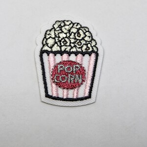 May include: White embroidered iron-on patch with a popcorn bucket design. The bucket is pink and white striped with a red circle that says "POP CORN" in silver lettering.