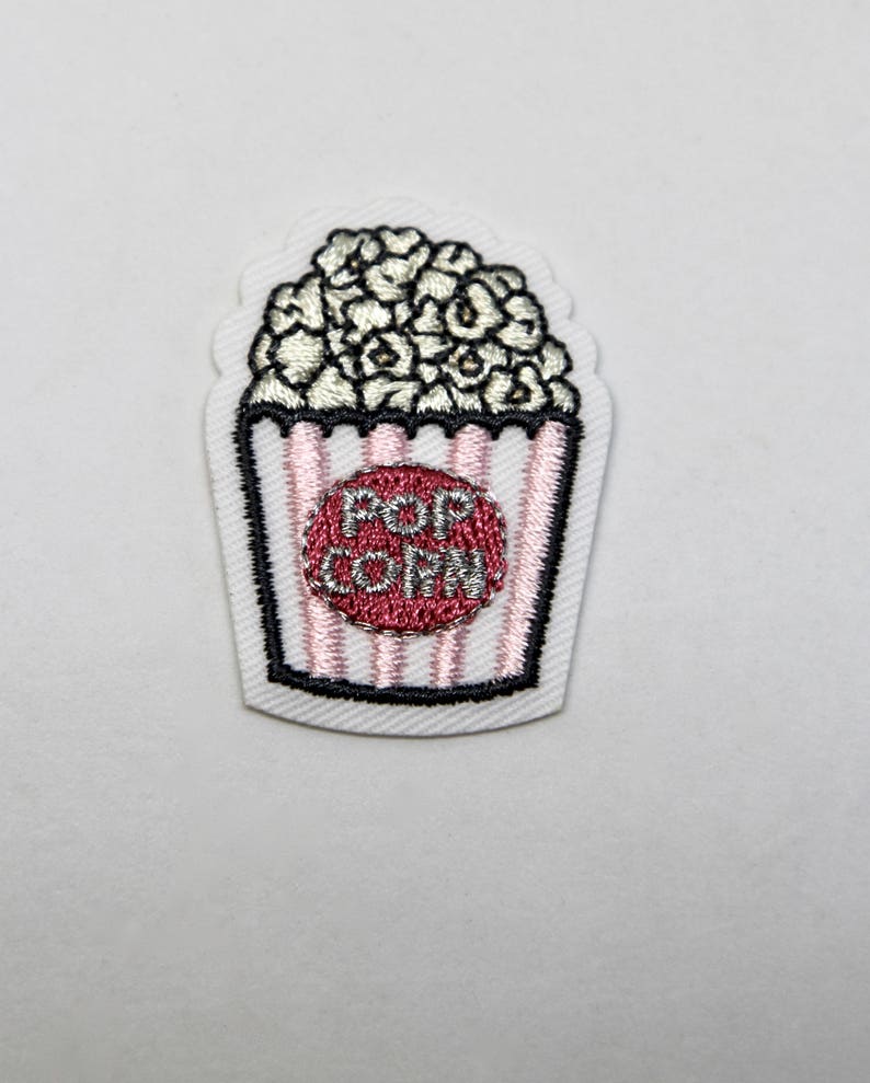 May include: White embroidered iron-on patch with a popcorn bucket design. The bucket is pink and white striped with a red circle that says "POP CORN" in silver lettering.