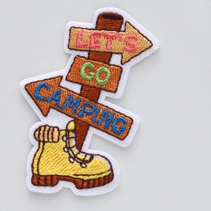 May include: Embroidered iron-on patch with a brown wooden signpost with three arrows pointing in different directions. The arrows have the text "Let's Go Camping" on them. A yellow hiking boot is at the bottom of the signpost.