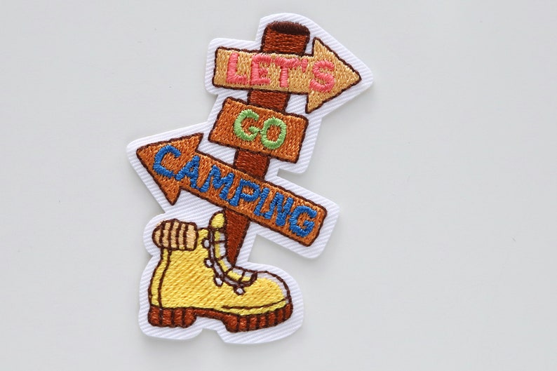 May include: Embroidered iron-on patch with a brown wooden signpost with three arrows pointing in different directions. The arrows have the text "Let's Go Camping" on them. A yellow hiking boot is at the bottom of the signpost.