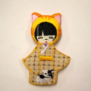 May include: Embroidered felt doll wearing a yellow cat costume and a brown and white patterned kimono. The doll is holding a small white dog with black spots.