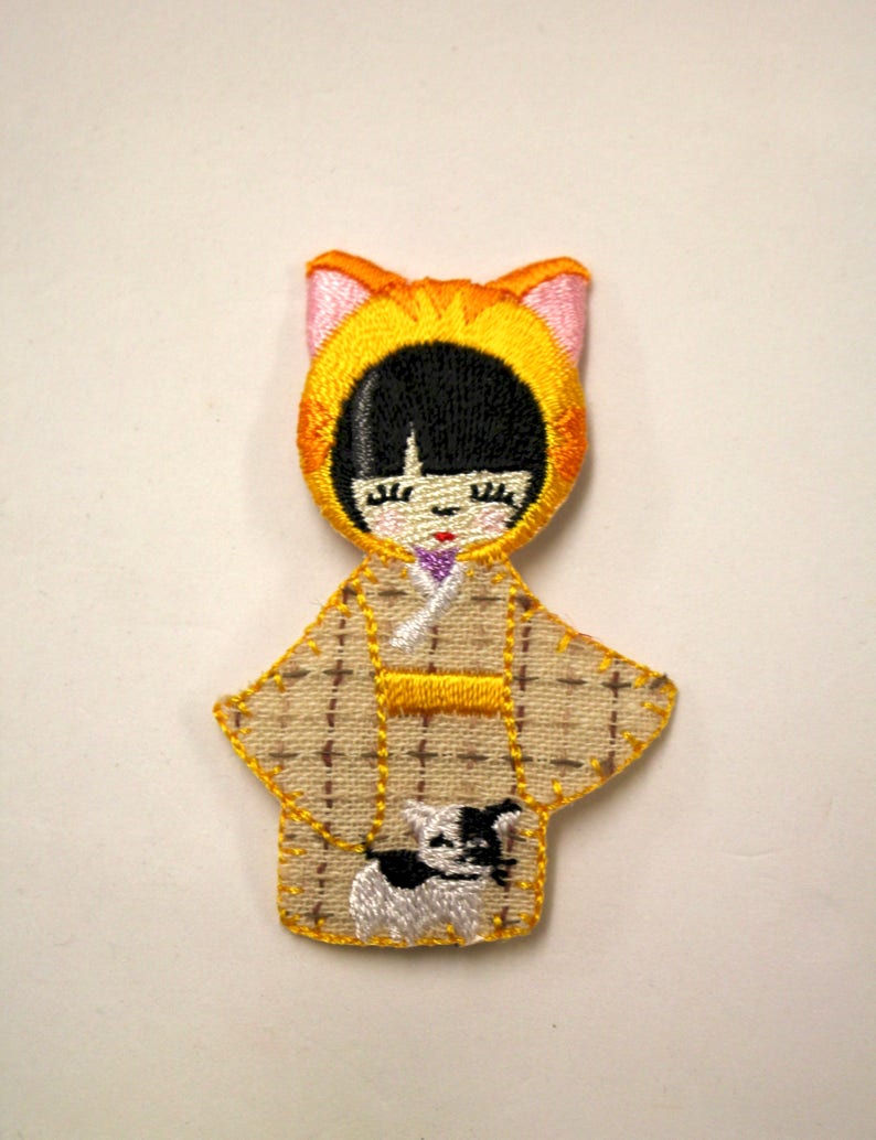 May include: Embroidered felt doll wearing a yellow cat costume and a brown and white patterned kimono. The doll is holding a small white dog with black spots.
