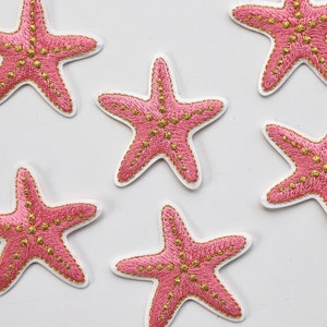 Puede incluir: Seis parches bordados de estrella de mar rosa y oro. Las estrellas de mar son de color rosa claro con costuras y detalles dorados.