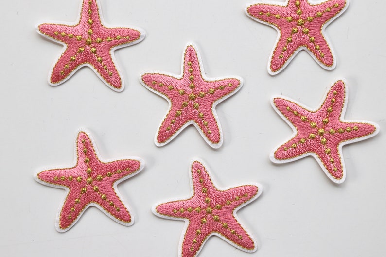 Puede incluir: Seis parches bordados de estrella de mar rosa y oro. Las estrellas de mar son de color rosa claro con costuras y detalles dorados.