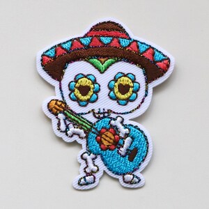 May include: Embroidered iron-on patch of a skeleton wearing a sombrero and playing a guitar. The skeleton has colourful flowers on its skull and is wearing a blue shirt.