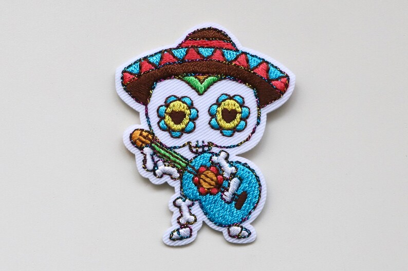 May include: Embroidered iron-on patch of a skeleton wearing a sombrero and playing a guitar. The skeleton has colourful flowers on its skull and is wearing a blue shirt.