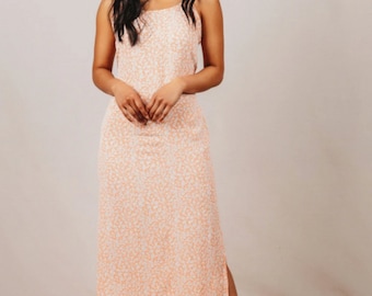 May include: A woman wearing a peach-coloured maxi dress with a white floral print. The dress has a spaghetti strap design and a side slit. She is wearing beige and white checkered slip-on shoes.