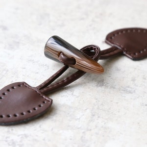 May include: A brown leather toggle closure with a horn toggle. The leather has stitching around the edges.