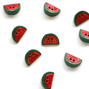 May include: A collection of 11 watermelon-shaped buttons in red and green. The buttons are arranged in a scattered pattern on a white background.