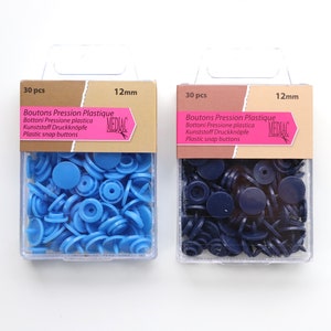 K&ouml;nnte beinhalten: Zwei Packungen mit 12 mm Kunststoff-Druckkn&ouml;pfen. Eine Packung enth&auml;lt 30 hellblaue Kn&ouml;pfe und die andere Packung enth&auml;lt 30 dunkelblaue Kn&ouml;pfe. Die Packungen sind mit "Boutons Pression Plastique" und "MEDIAC" beschriftet.