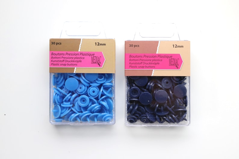 K&ouml;nnte beinhalten: Zwei Packungen mit 12 mm Kunststoff-Druckkn&ouml;pfen. Eine Packung enth&auml;lt 30 hellblaue Kn&ouml;pfe und die andere Packung enth&auml;lt 30 dunkelblaue Kn&ouml;pfe. Die Packungen sind mit "Boutons Pression Plastique" und "MEDIAC" beschriftet.